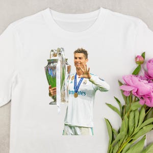 May include: White t-shirt featuring a graphic of a soccer player holding a trophy and waving. The player is wearing a white jersey with blue accents and the word "emirates" printed on the front. A gold medal hangs around his neck.