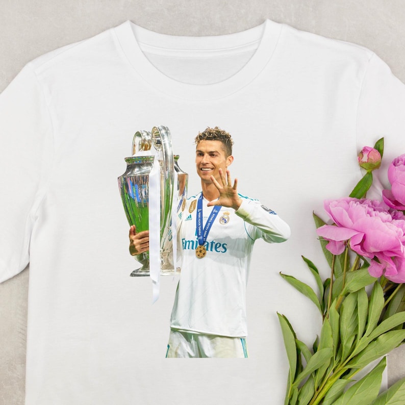 May include: White t-shirt featuring a graphic of a soccer player holding a trophy and waving. The player is wearing a white jersey with blue accents and the word "emirates" printed on the front. A gold medal hangs around his neck.