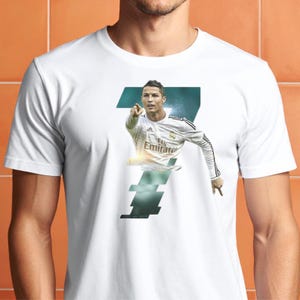 May include: White t-shirt featuring a graphic of a soccer player pointing, with the number 7 in the background. The player is wearing a white jersey with the Emirates logo. The shirt is a classic crew neck style.