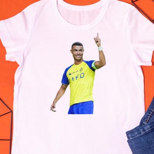 May include: White t-shirt featuring a vibrant image of a soccer player in a yellow and blue uniform, pointing upwards. The shirt showcases a detailed graphic of the athlete, perfect for sports fans.