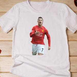 May include: White t-shirt featuring a graphic of a soccer player in a red jersey with the number 7. The player is in a celebratory pose, with his mouth open and arm raised. The jersey has the letters "IG" on it.
