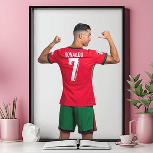 May include: Framed print of a soccer player in a red jersey with the name "RONALDO" and the number "7" on the back. The player is flexing his arms, wearing green shorts. The image is set against a white background.