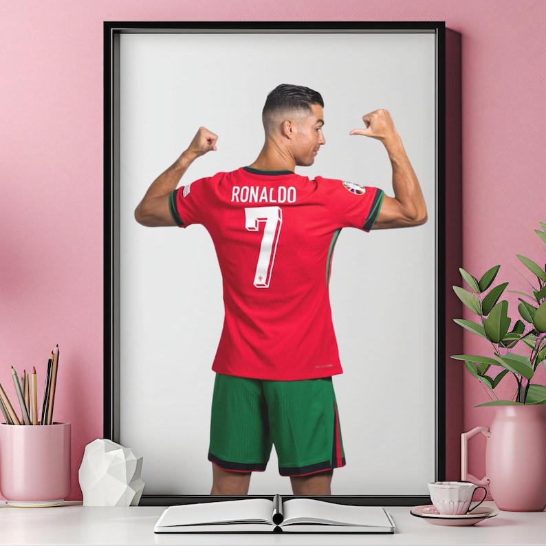 May include: Framed print of a soccer player in a red jersey with the name "RONALDO" and the number "7" on the back. The player is flexing his arms, wearing green shorts. The image is set against a white background.