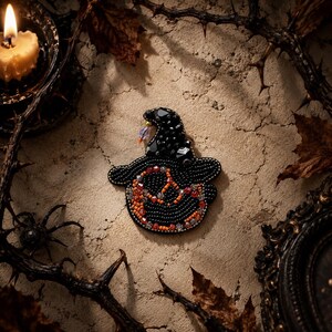 May include: A handcrafted brooch shaped like a witch hat, featuring black beads, faceted black crystals, and a band of orange and clear beads. The design includes a small dangling purple crystal accent at the tip of the pointed hat structure.