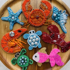 Peut inclure: Collection d'animaux marins perlés, dont une étoile de mer, un crabe, une crevette, une tortue, un homard et un poisson. Les créatures sont de différentes couleurs, notamment orange, bleu, rouge, vert et rose, sur une surface en bois.