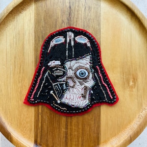May include: Embroidered patch featuring a stylized depiction of Darth Vader's helmet, with a skeletal face and a single blue eye. The patch is outlined in red and black, with intricate beadwork details.