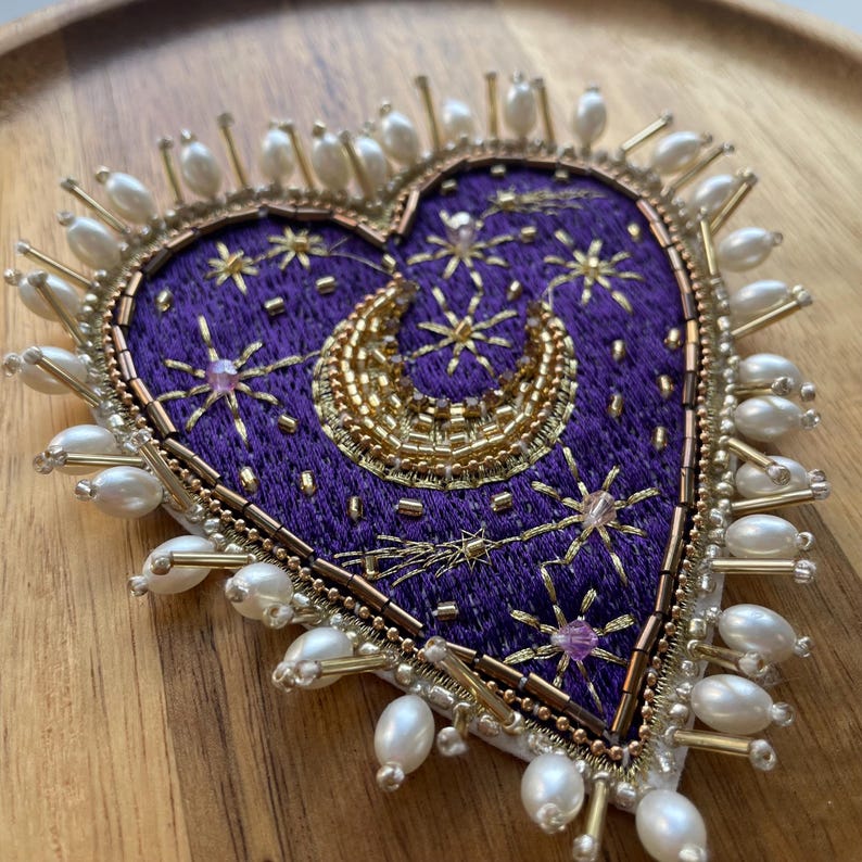Pu&ograve; includere: Spilla a forma di cuore con sfondo ricamato viola, ornata da abbellimenti color oro e perle finte. Il design include una luna crescente e motivi a stella. La spilla &egrave; larga circa 7.6 cm.