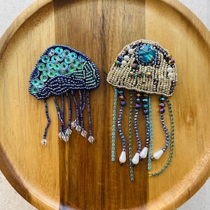 May include: Two handmade jellyfish brooches. One is navy blue with teal sequins and beaded tentacles. The other is gold and teal with a large teal gem and dangling beaded strands. Both are on a wooden tray.