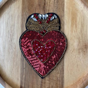 May include: A heart-shaped patch with a red sequined design and a smaller heart in the center. The patch is outlined with gold beads and has a black backing. The top features gold beading and red and clear crystal accents.