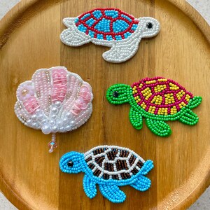 Könnte beinhalten: Drei perlenbesetzte Schildkröten-Aufnäher und ein perlenbesetzter Muschel-Aufnäher. Die obere Schildkröte ist weiß, blau und rot. Die mittlere Schildkröte ist grün, gelb und rosa. Die untere Schildkröte ist blau, weiß und braun. Die Muschel ist rosa und weiß mit einer hängenden Perle.
