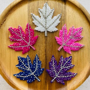 Könnte beinhalten: Fünf aufgenähte Ahornblätter aus Perlen in Weiß, Rosa, Blau und Lila. Jedes Blatt ist mit glitzernden Strasssteinen entlang der Adern verziert. Die Applikationen sind auf einem Holztablett angeordnet und zeigen ihre leuchtenden Farben und filigranen Details.