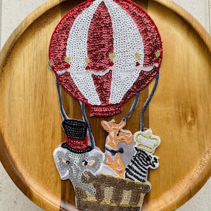 Könnte beinhalten: Ein skurriles Pailletten-Patch mit einem rot-weiß gestreiften Heißluftballon. Darunter befindet sich ein Korb mit einem Elefanten mit Zylinder, einer Giraffe und einem Zebra mit Krone. Ideal zum Aufnähen auf Kleidung oder für Bastelprojekte.