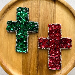 Può includere: Due toppe a forma di croce, una ricoperta di paillettes verde smeraldo e l'altra di paillettes rosso rubino. Ogni croce è delineata da un bordo di piccoli strass trasparenti e riflettenti, creando un contorno scintillante.