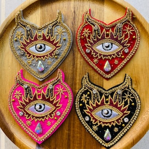 May include: Four heart-shaped embroidered patches in gray, red, pink, and black. Each features an eye design with gold and red accents, a teardrop gem, and star details. The patches are embellished with beads and sequins.