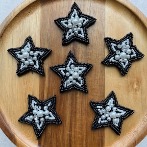 May include: Six black star-shaped beaded embellishments with white and pearl-like beads. The stars are arranged on a wooden surface. The stars have a beaded outline and are filled with various sizes of white and pearl-like beads.