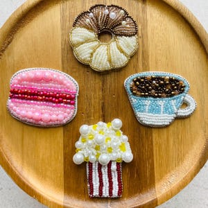 Pode incluir: Quatro broches de missangas em forma de alimentos: um donut castanho e branco, um macaron rosa e vermelho, uma chávena de café azul-clara e branca cheia de missangas castanhas e um balde de pipocas às riscas brancas e vermelhas.