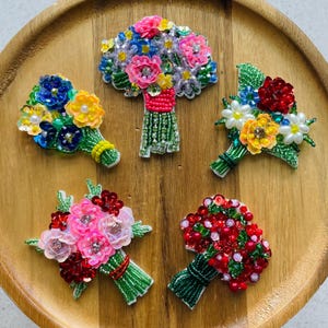 Op de afbeelding: Vijf handgemaakte bloemenbroches met ingewikkeld kralen- en paillettenwerk. Elk stuk toont een boeket met levendige kleuren, waaronder roze, rood, blauw, geel en groen. De ontwerpen tonen gedetailleerde stelen en bloemblaadjes gemaakt met kleine glaskralen, pailletten en centrale kristallen accenten, gerangschikt in verschillende clusters.
