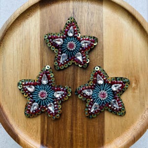 May include: Three star-shaped decorative patches. Each star features a teal center with a pink center stone, surrounded by clear, teardrop-shaped crystals, red beads, and gold sequins. The patches are on a wooden surface.