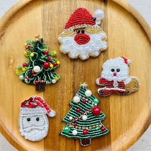 Può includere: Cinque spille di perline fatte a mano con temi natalizi. Due spille sono alberi di Natale verdi decorati con perline rosse, bianche e gialle. Tre spille raffigurano figure di Babbo Natale in vari stili, realizzate con perline di vetro rosse, bianche e nere.