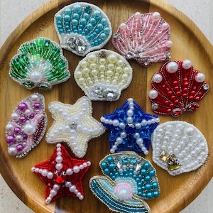 以下が含まれることがあります： 青、ピンク、緑、赤、白など、さまざまな色のビーズで飾られた貝殻とヒトデの装飾品のコレクション。 各作品はパールとラインストーンで飾られ、きらめく効果を生み出しています。 木のトレイに配置されています。