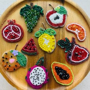 May include: A collection of ten fruit-shaped beaded pins featuring a pomegranate, grapes, apple, orange slice, lemon, watermelon slice, orange, fig, dragon fruit, and papaya, all crafted with colorful glass beads and sequins on a wooden tray.