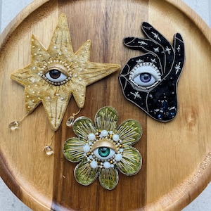May include: Three embroidered eye-themed brooches. A gold star with a brown-eyed eye, a black hand with a blue-eyed eye, and a green flower with a green-eyed eye. Each brooch is embellished with beads and crystals.