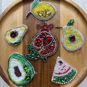 Puede incluir: Una colección de broches con forma de fruta con cuentas, que incluyen un limón, un aguacate, una granada y una sandía. Los broches presentan colores vibrantes, intrincados abalorios y detalles de piedras preciosas. Los broches se exhiben en una bandeja de madera.