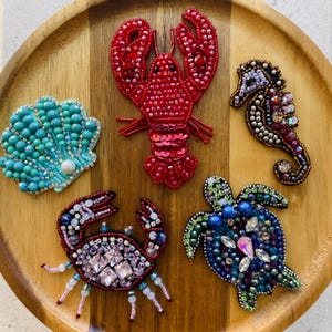 May include: A collection of beaded sea creature brooches, including a red lobster, a turquoise seashell, a seahorse, a crab, and a sea turtle. The brooches are embellished with sequins and crystals, set on a wooden tray.