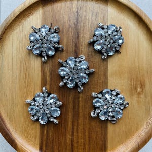 May include: Five silver-toned flower-shaped embellishments with clear, faceted crystal accents. Each has a central cluster of smaller crystals and larger, teardrop-shaped crystals around the perimeter. They are arranged on a wooden surface.