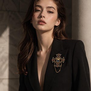 May include: A gold-toned brooch shaped like an ornate cross featuring a central blue eye motif, intricate filigree detailing, small red accents, and a delicate hanging chain, pinned to the lapel of a structured black blazer.