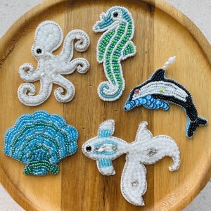 Könnte beinhalten: Fünf handgefertigte Perlenaufnäher mit Meeresmotiven: ein weißer Oktopus, ein grün-blaues Seepferdchen, ein schwarz-blauer Delfin, eine blau-grüne Muschel und ein weißer Stachelrochen mit blauen Akzenten, alle mit kleinen Glasperlen verziert.