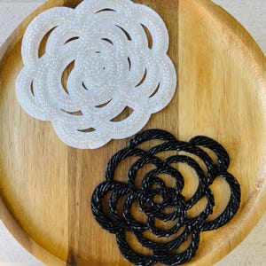 Pode incluir: Dois enfeites em forma de flor. Um é branco com uma textura de contas, e o outro é preto com uma textura de contas. Ambos estão em uma bandeja de madeira. A flor branca tem uma borda ondulada e um centro em espiral. A flor preta tem um design semelhante.