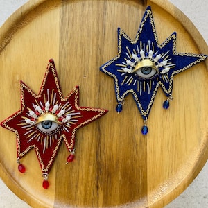 May include: Two star-shaped embroidered patches, one in red velvet and one in blue velvet. Each features a central eye motif with gold-tone metal eyelids, surrounded by radiating metallic threads, white beads, and dangling crystal-like drops.