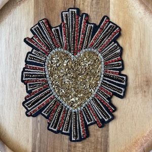 Puede incluir: Un parche en forma de corazón con un centro de lentejuelas doradas y un borde negro. El parche presenta rayos de cuentas rojas y doradas que emanan del corazón, creando un efecto de sol. El parche está sobre una superficie de madera.