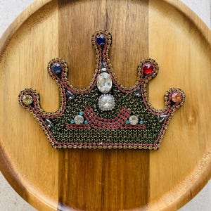 May include: A decorative crown with a black base, embellished with pink, green, and gold beading. The crown features various colourful gemstones, including red, blue, and clear crystals. The crown is set on a wooden surface.