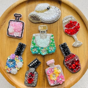 May include: A collection of eight handcrafted beaded pins featuring various shapes including a white sun hat, a wine glass filled with red beads, and several perfume bottles decorated with colorful floral accents, sequins, and metallic beads.