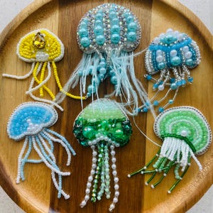 May include: A collection of handmade beaded jellyfish brooches in various colors, including yellow, blue, green, and white. Each brooch features intricate beadwork, pearl accents, and delicate tendrils, displayed on a wooden tray.
