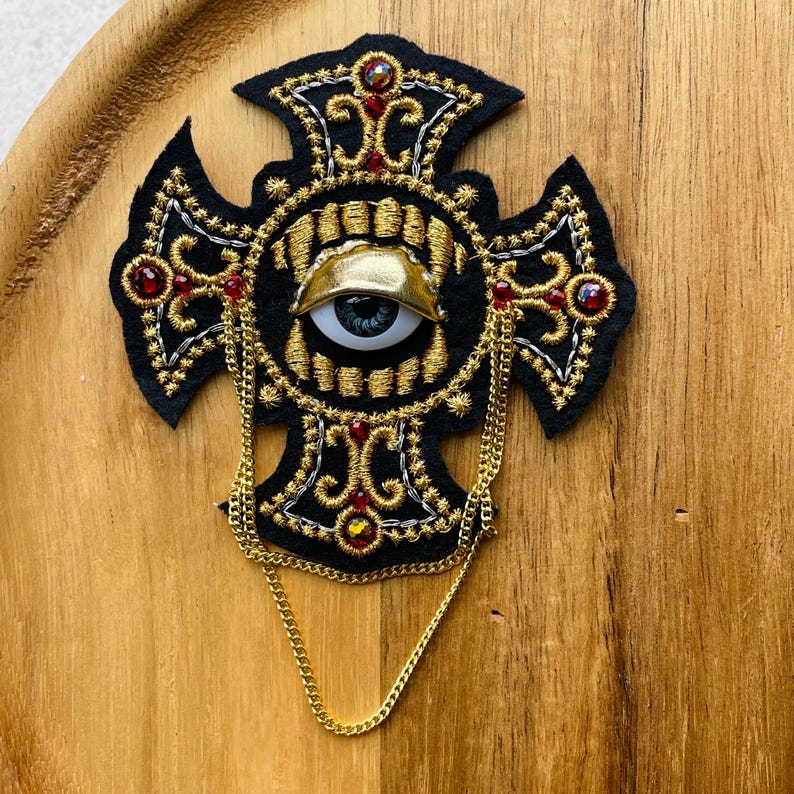 May include: A black felt cross brooch featuring intricate gold and silver embroidery, a central realistic eye with a gold eyelid, and small red crystals. A thin gold-toned metal chain hangs from the sides of the cross, adding a layered element to the detailed, textured design of this decorative accessory.