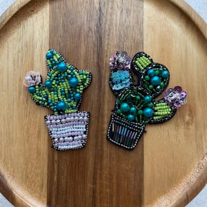 Könnte beinhalten: Zwei bestickte Kaktus-Anstecknadeln. Ein Kaktus ist grün und türkis mit einer rosa Blüte, der andere ist grün und türkis mit rosa und blauen Blüten. Beide befinden sich in dekorativen Töpfen mit Perlen- und Pailletten-Details.