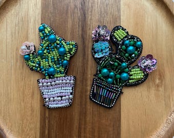 May include: Two beaded cactus pins. One cactus is green and turquoise with a pink flower, the other is green and turquoise with pink and blue flowers. Both are in decorative pots with pearl and sequin details.