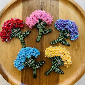 May include: Five beaded flower brooches in red, pink, purple, blue, and yellow. Each brooch features a cluster of sequins, green beaded stems, and leaves. The brooches are arranged on a wooden tray.