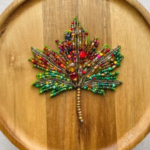 Pode incluir: Um broche em forma de folha de bordo feito de feltro verde, densamente adornado com uma variedade vibrante de contas de vidro multicoloridas, contas metálicas tubulares e cristais facetados. O caule é coberto por pequenas contas douradas.