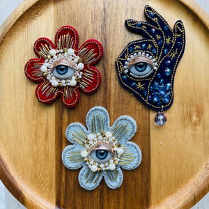 May include: Three handmade brooches: a red flower, a blue hand, and a light blue flower. Each features a blue eye, gold and white bead accents, and intricate embroidery. The hand brooch has a dangling bead.