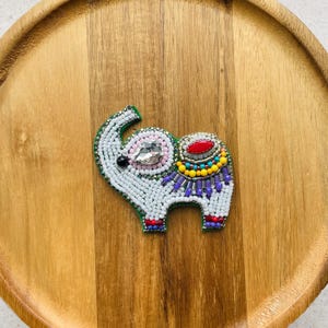 May include: An elephant-shaped brooch crafted with white seed beads, featuring a clear crystal eye, a red oval cabochon on the back, and decorative accents in yellow, turquoise, and purple beads. The edges are finished with green thread.