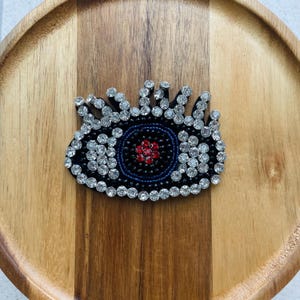 May include: A black eye-shaped brooch with a blue and red centre, surrounded by sparkling clear crystals. The brooch is set against a wooden background, showcasing its intricate design. This accessory adds a touch of glamour and style.