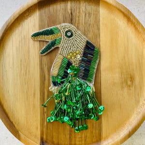 Könnte beinhalten: Eine toucanförmige Brosche aus detaillierten Perlenstickereien in Gold, Grün und Dunkellila. Der Vogel hat einen markanten Schnabel, ein einzelnes Kristallauge und einen herabhängenden Fransenbesatz aus hellgrünen Pailletten und Perlen.