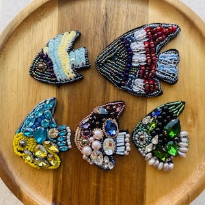 May include: Six beaded fish brooches in a range of colours, including blue, yellow, red, and green. Each brooch has detailed beadwork and embellishments, with different shapes and sizes. The brooches are presented on a wooden tray.
