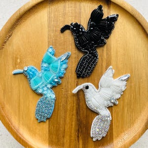 May include: Three hummingbird-shaped brooches crafted with beads, sequins, and crystals. One brooch is solid black, one is bright turquoise blue, and one is white with iridescent accents. Each bird is depicted in flight with wings spread.