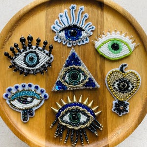 Könnte beinhalten: Eine Sammlung von sieben bestickten Aufnähern mit Augenmotiven in verschiedenen Formen und Farben. Die Designs enthalten Perlen, Pailletten und metallische Akzente in Blau, Grün, Schwarz, Weiß und Gold, angeordnet auf einer runden Holzoberfläche.