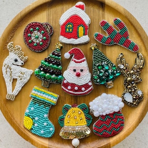 Könnte beinhalten: Eine Sammlung von elf perlenbesetzten Anstecknadeln mit Weihnachtsmotiven, darunter ein weißes Rentier, ein rotes Haus, ein grüner Kaktus, eine Kugel, zwei Weihnachtsbäume, ein Weihnachtsmann, ein blauer Strumpf, eine gelbe Glocke und ein goldener Hirsch.